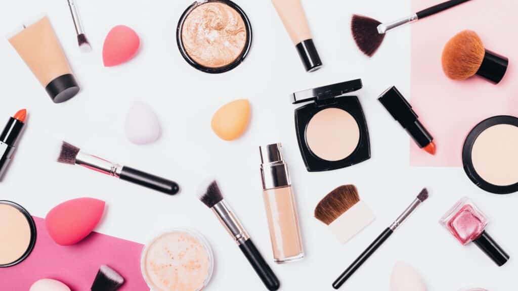 an assortment of beauty brand products lay on a white clear table with makeup brushes lipstick and moisturizers